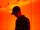 Ein Junge mit Basecap und Brille steht vor einem orangenen Vorhang und schaut traurig auf den Boden