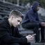 Zwei Teenager sitzen an einem düsteren Tag auf einer leeren Sporttribüne. Ein Teenager schaut gedankenverloren aufs Smartphone. 