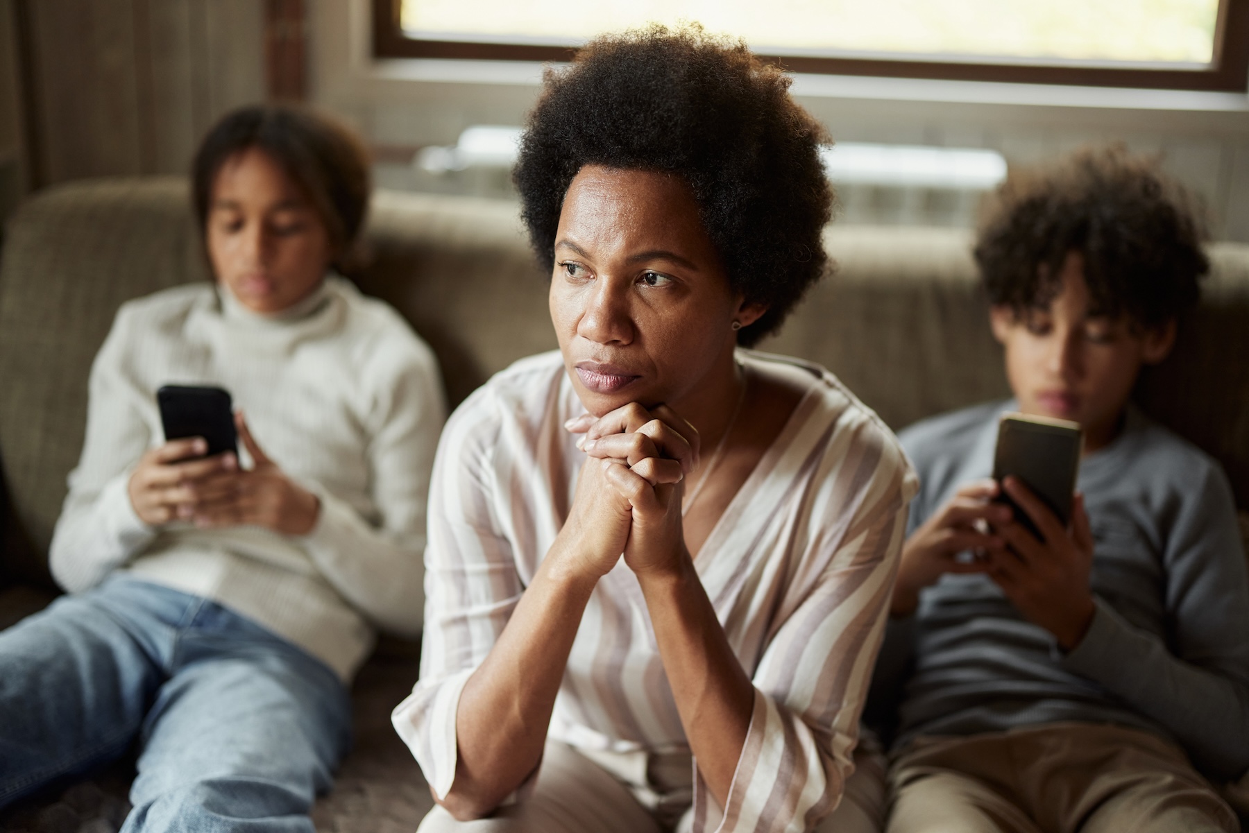 Mutter sitzt nach vorne gelehnt auf dem Sofa und schaut nachdenklich, neben ihr sitzen zwei Jungen mit ihren Smatphones