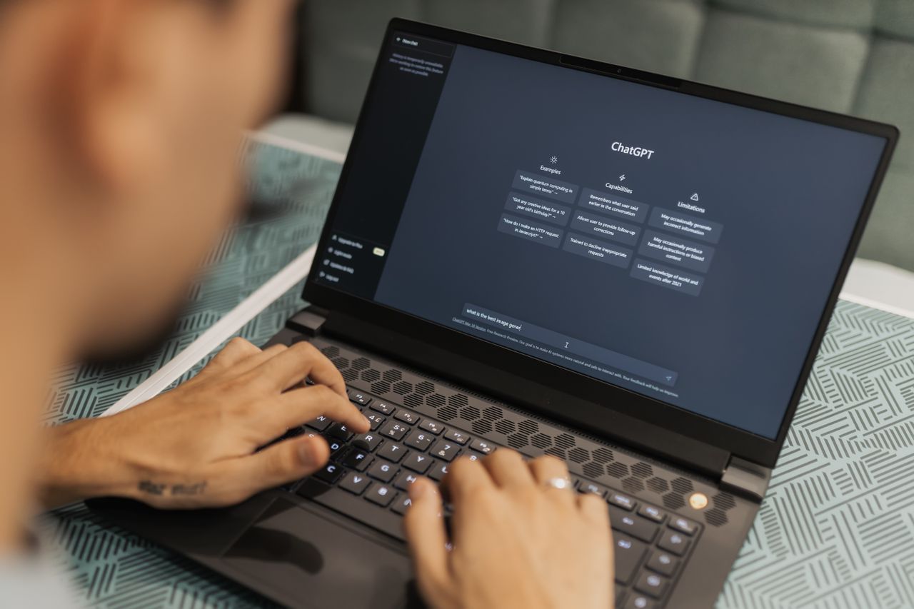 Eine Person sitzt an einem Tisch und tippt auf der Tastatur eines Laptops. Auf dem Bildschirm ist die ChatGPT-Oberfläche geöffnet.