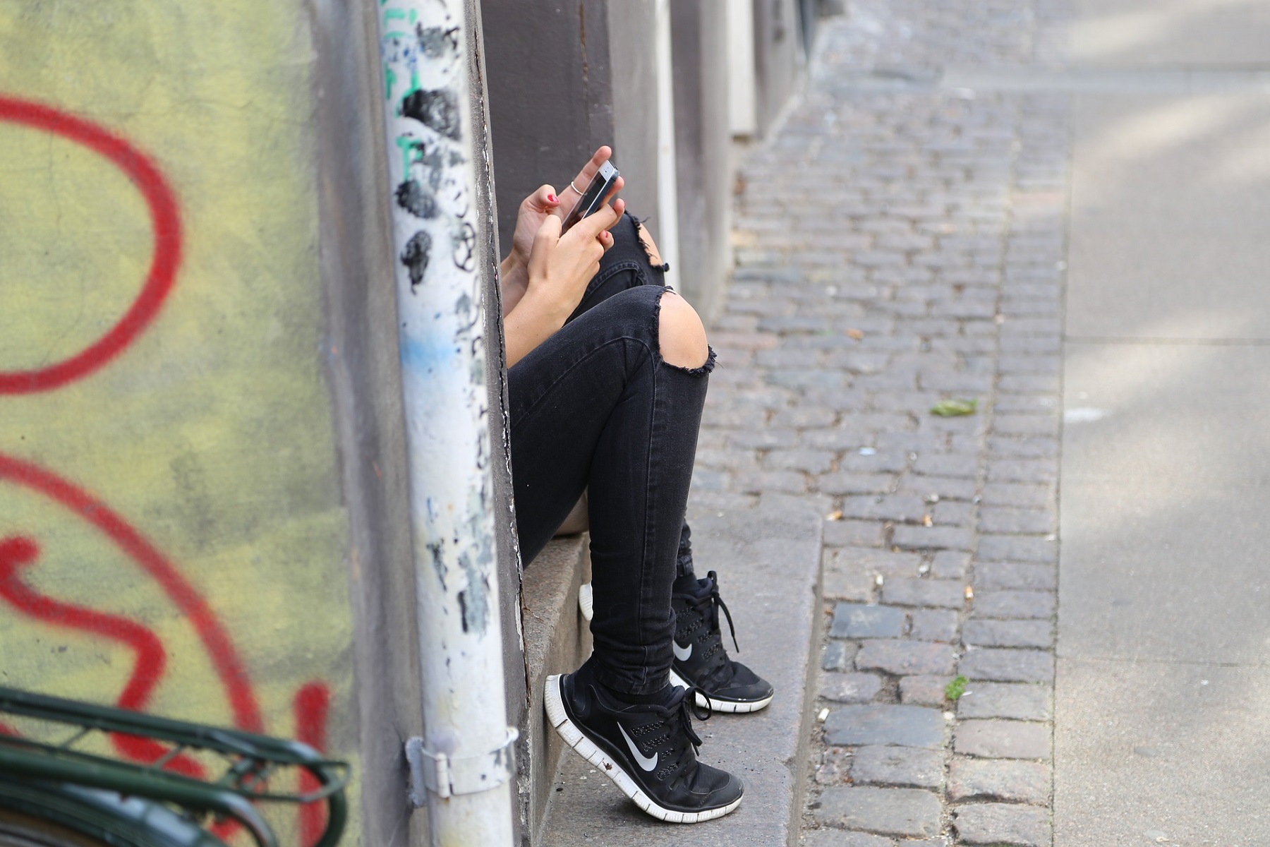 hinter einer Wand mit Graffiti sitzt eine jugendliche Person, von der man nur die Beine in zerrissenen Jeans und Sneakers und die Hände mit einem Smartphone sieht.
