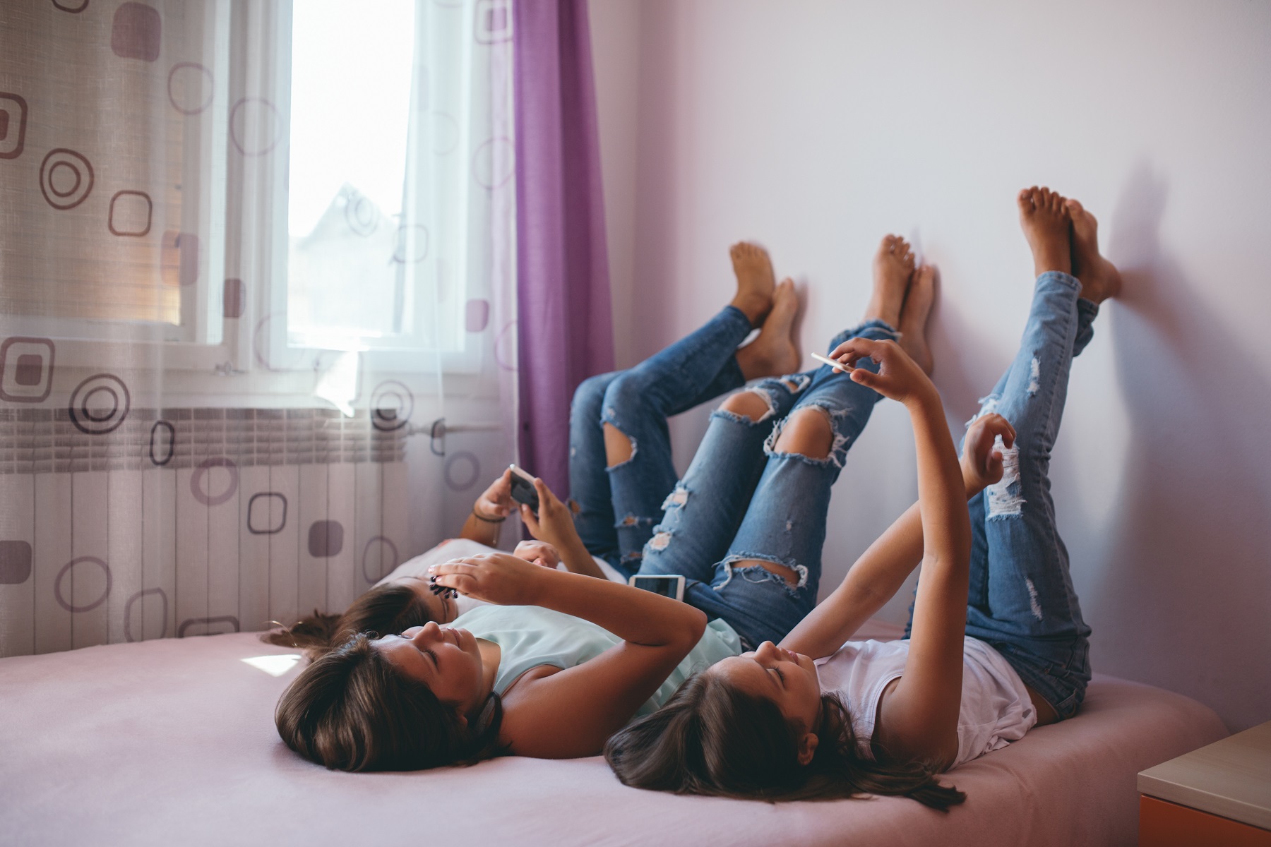 Drei Mädchen liegen nebeneinander auf dem Bett mit den Füßen an der Wand und Smartphones in der Hand