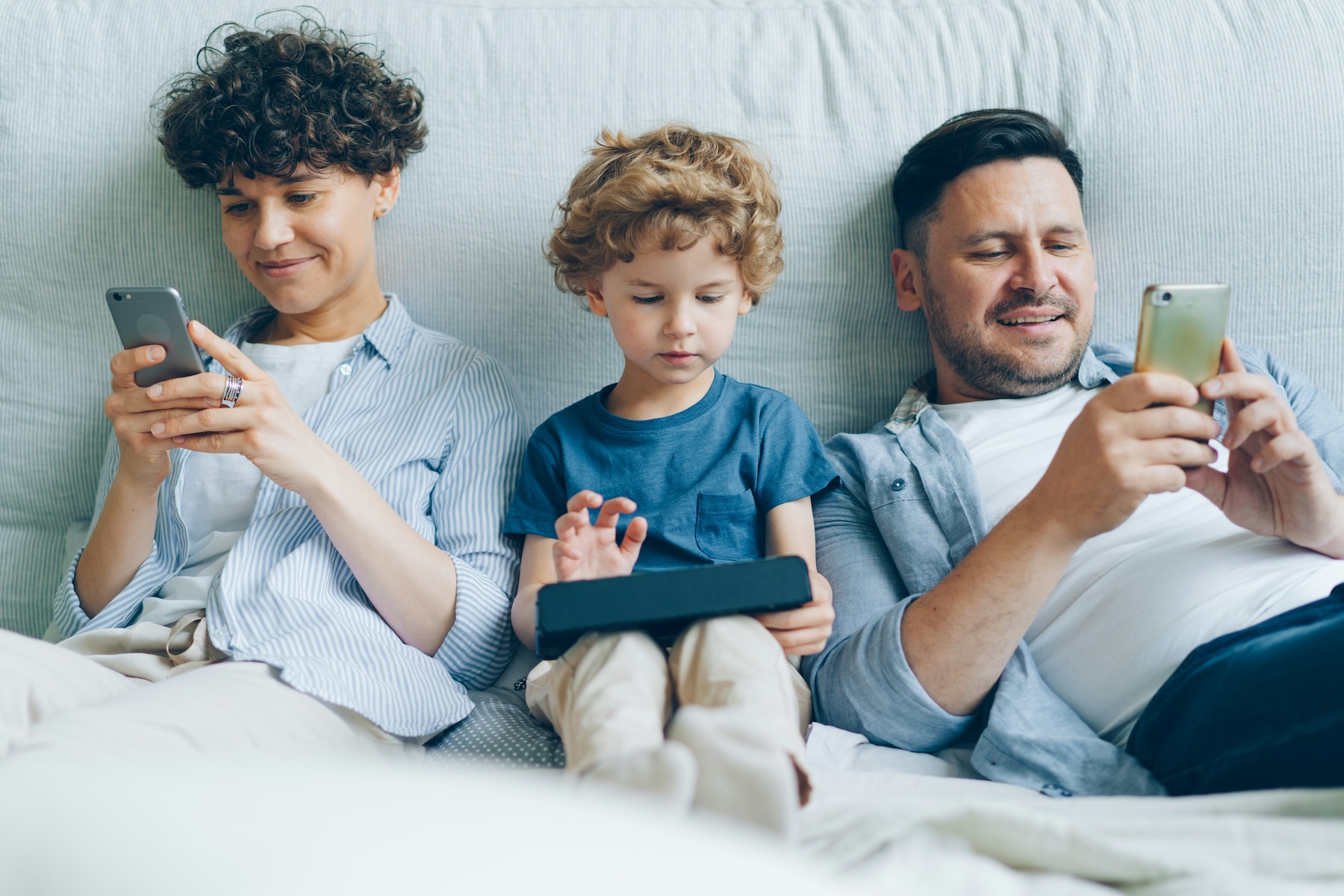Mutter, Sohn und Vater liegen entspannt nebeneinander im Bett. Alle schauen auf ein eigenes Smartphone.