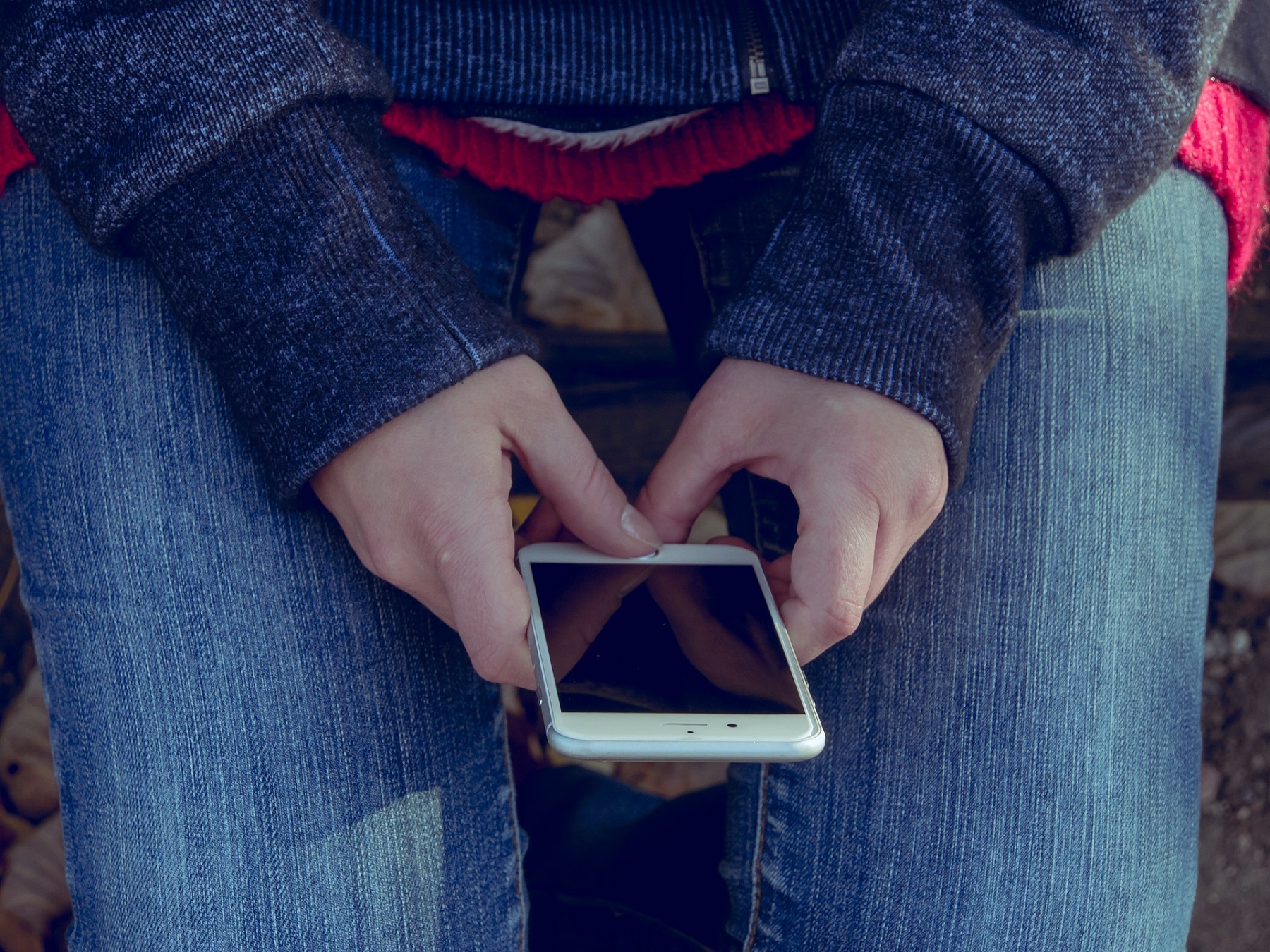 Person mit blauem Pulli und Jeans hält ein Smartphone in der Hand