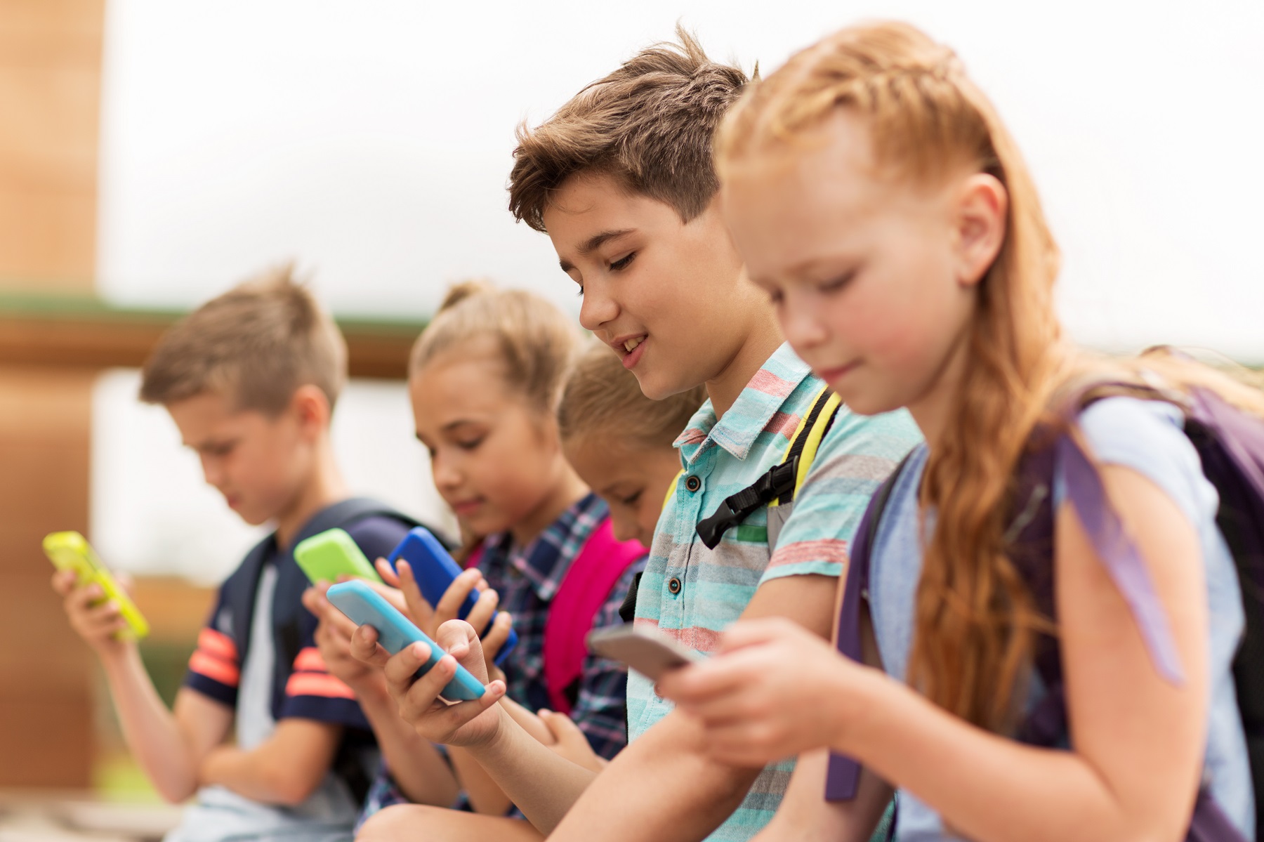 Kinder mit Schulranzen stehen nebeneinander und schauen auf ihre bunten Smartphones.