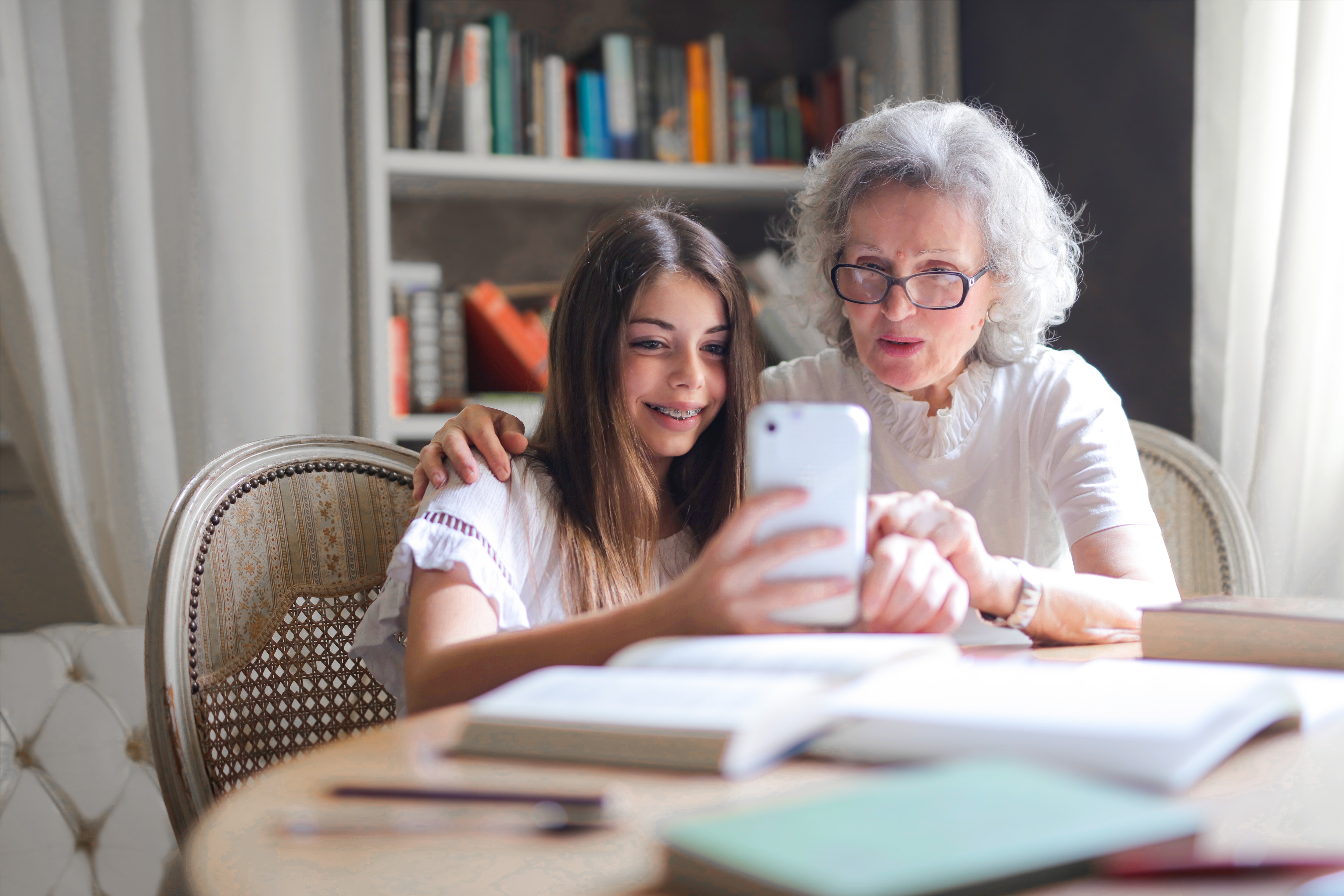 Eine grauhaarige Dame auf der rechten Seite sitzt neben einem Mädchen mit braunen Haaren. Sie blicken beide auf ein weißes Smartphone in Richtung Kamera.