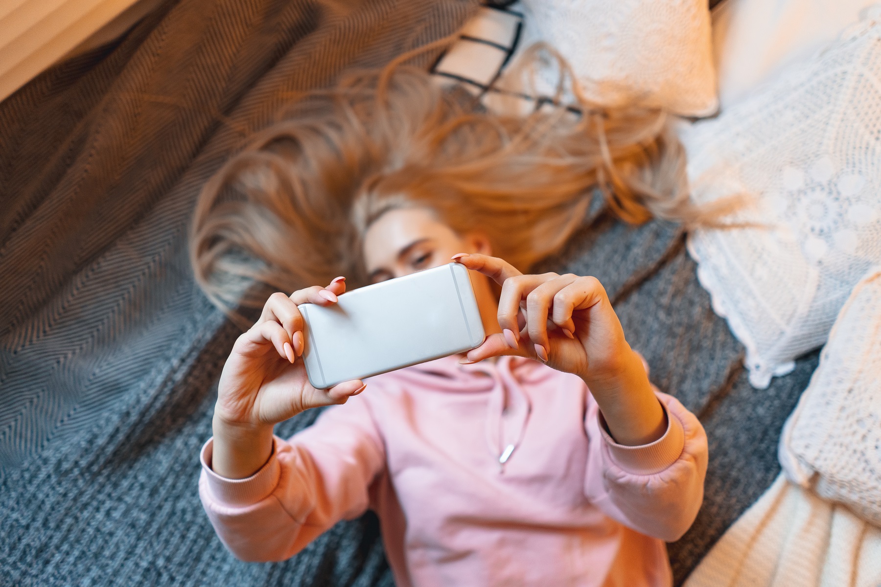 Junge Frau mit blonden Haaren liegt auf dem Boden und hält ihr Smartphone in die Höhe.