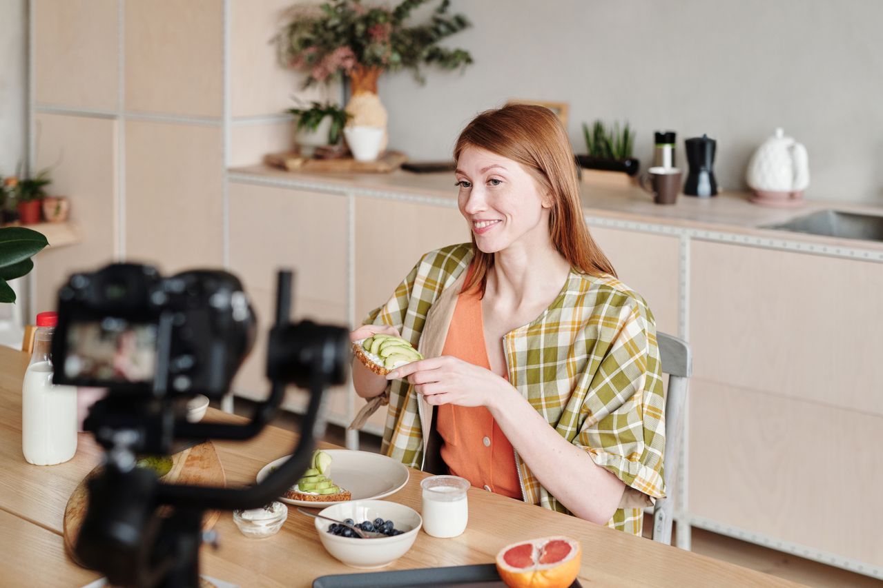Eine Frau sitzt an einem Tisch, vor ihr eine Kamera auf einem Stativ. Sie hält ihr Avocado-Brot in die Kamera.