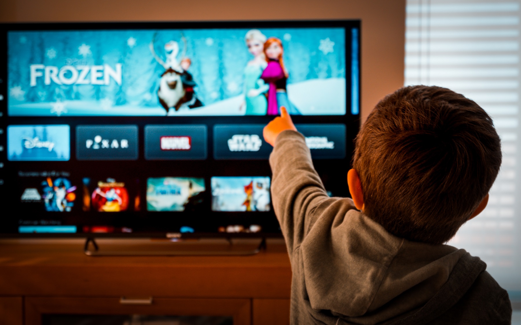 Von hinten ist ein Junge zu sehen, der auf einen Fernseher zeigt, auf dem der Streaming-Dienst "Disney Plus" läuft