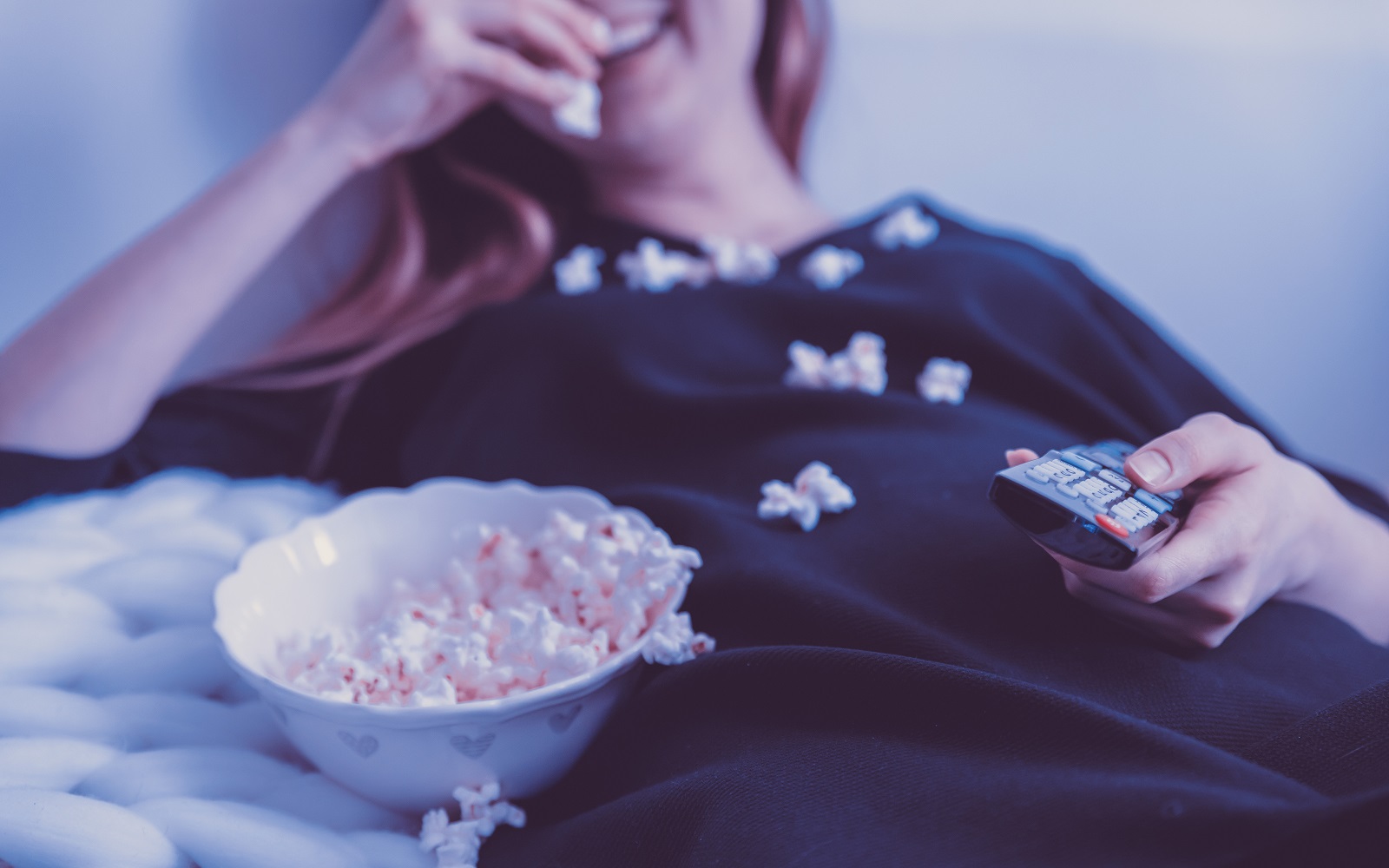 Eine junge Frau isst lachend Popcorn und hält eine Fernbedienung in der Hand