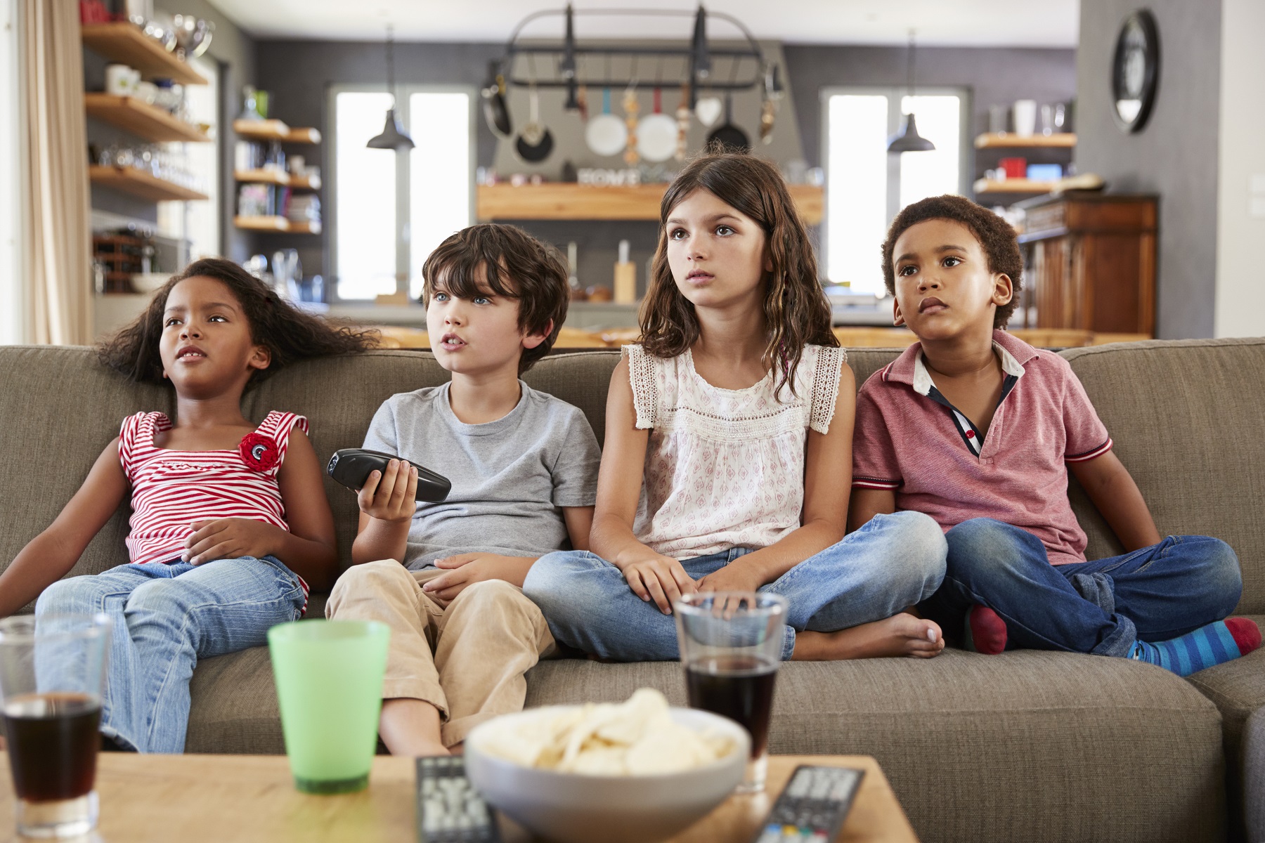 Vier Kinder sitzen auf der Couch, einer von ihnen hat die Fernbedienung in der Hand und sie sehen alle in die selbe Richtung.